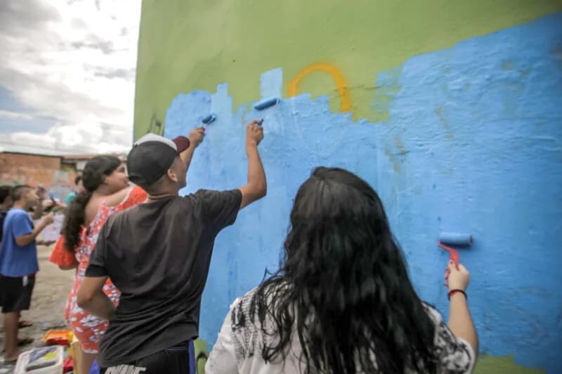 Jovens com rolos de tinta pintam de azul a parede externa de um prédio, case HOUSECULTURA