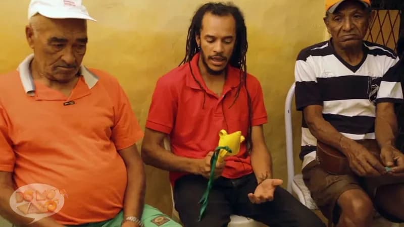 Três homens sentados conversam, um deles com uma flor amarela de pano na mão, websérie DôMóValô da HOUSECULTURA