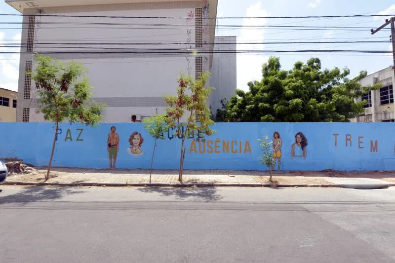 Muro azul com figuras pintadas e as palavras PAZ e AUSÊNCIA em calçada arborizada, case HOUSECULTURA