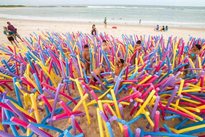 Instalação de macarrões de piscina coloridos fincados na areia com crianças brincando, festival Arte Praia, case HOUSECULTURA