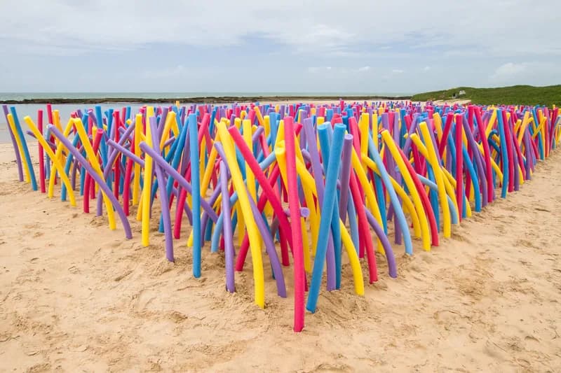 Faixa contínua de macarrões de piscina coloridos fincados na areia junto ao mar, festival Arte Praia, case HOUSECULTURA