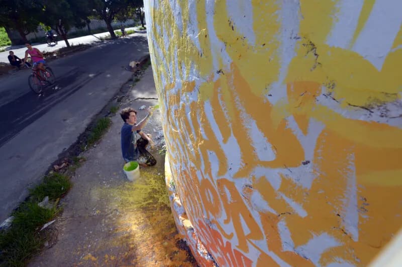 Rapaz pinta a base de um muro amarelo e branco na rua enquanto um ciclista passa, case HOUSECULTURA