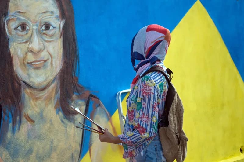 Artista com lenço na cabeça segura pincéis diante de mural com o retrato de uma senhora, case HOUSECULTURA