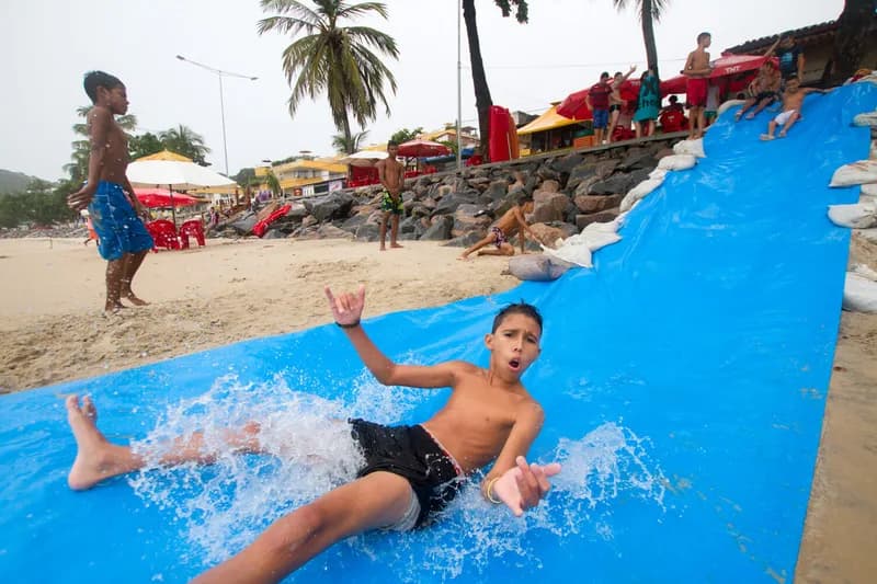 Menino escorrega por lona azul molhada na areia durante o festival Arte Praia, case HOUSECULTURA