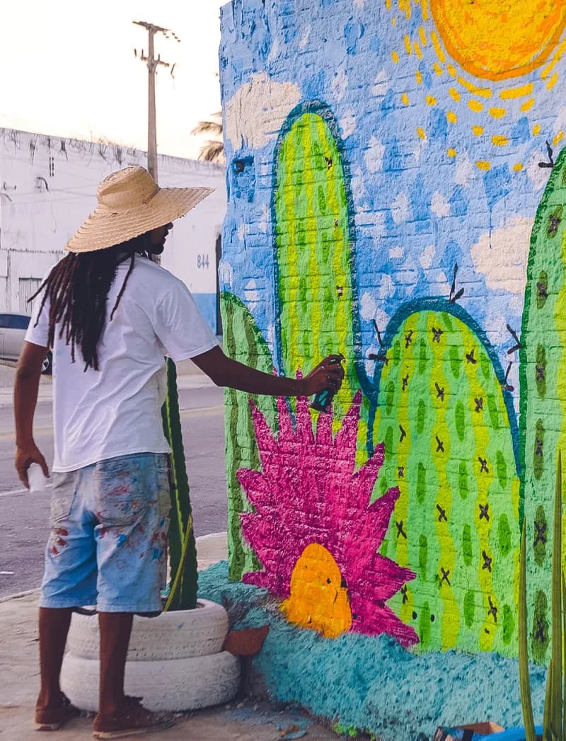 Artista de chapéu de palha pinta mural de cactos e sol em muro de rua de Fortaleza, case HOUSECULTURA